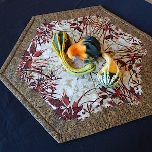 May include: Three gourds in a variety of colours and shapes on a brown and gold patterned fabric. The gourds are green, orange, and black.