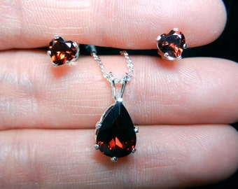 May include: A jewellery set comprising a silver necklace with a teardrop-shaped, dark red gemstone pendant, and matching heart-shaped stud earrings. The gemstones have a faceted appearance. The set is displayed against a dark background.