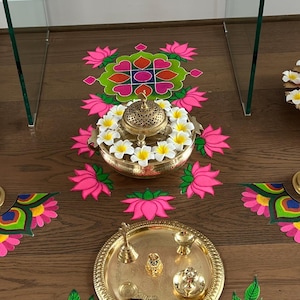 May include: A black statue of Ganesha sits atop a white table decorated with pink floral designs. The table is made of glass and has a gold-colored base. The floor is decorated with colorful floral and peacock designs.