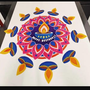 May include: A colorful rangoli design on a white surface featuring a central blue diya with a yellow flame and the text HAPPY DEEPAVALI. The pattern includes a circular arrangement of pink and blue lotus-like petals surrounded by smaller diya motifs.