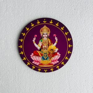 May include: Round decorative image featuring a seated deity, adorned with gold jewelry and holding flowers and a pot of gold coins. The image has a purple background and is surrounded by a gold border with small flame icons.