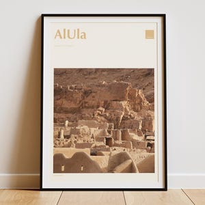 May include: Framed art print featuring the word "AlUla" in gold lettering above a photograph of an ancient desert city. The photograph shows a complex of tan-colored buildings nestled against a rocky cliff face.