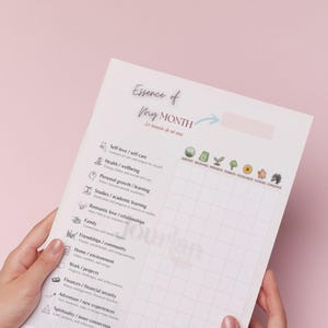 Puede incluir: Una página de diario blanca titulada "Essence of My Month" con una cuadrícula para el seguimiento de objetivos. La página incluye secciones para el amor propio, la salud, el crecimiento personal y más. El diario está sostenido por manos sobre un fondo rosa.