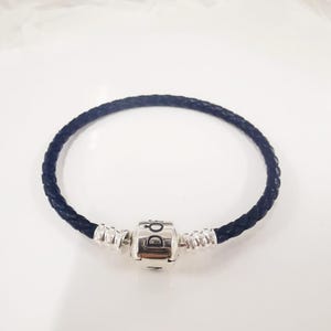 Op de afbeelding: Een donkerblauwe gevlochten leren armband met een zilverkleurige sluiting. De sluiting heeft een cilindrische vorm met de merknaam gegraveerd. De armband is een eenvoudig, elegant sieraad.