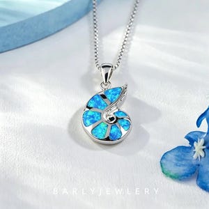 May include: A silver-toned necklace featuring a nautilus shell-shaped pendant inlaid with vibrant blue opal segments. The pendant hangs from a delicate box chain, and the text BARLYJEWLERY is printed in white at the bottom of the frame.