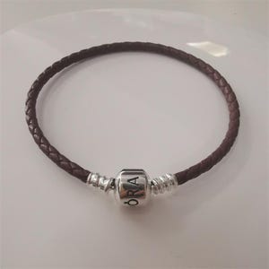 May include: A dark brown, braided leather bracelet with a silver-coloured barrel charm. The charm is engraved with the word "ORA". The bracelet is circular and rests on a white surface.