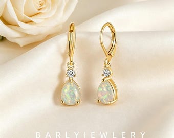 Pode incluir: Um par de brincos de ouro com fecho de alavanca, apresentando uma pedra de opala em forma de lágrima encimada por uma pequena pedra preciosa transparente de corte redondo. Os brincos estão sobre um tecido macio com uma rosa branca.