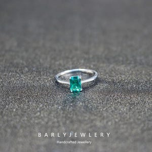 Peut inclure: Bague en argent avec une pierre verte rectangulaire taille émeraude. La bague est posée sur une surface texturée gris foncé. Le texte "BARLY JEWELRY Handcrafted Jewellery" est en bas.