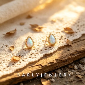 May include: A pair of gold stud earrings featuring teardrop-shaped opals with iridescent white and blue flashes. Each opal is encased in a delicate twisted rope-style gold metal border. The text BARLYJEWELRY is visible at the bottom of the image.