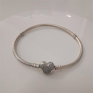 May include: A silver bracelet featuring a heart-shaped charm, studded with small, clear stones. The bracelet has a snake chain design and a clasp. A piece of jewellery.