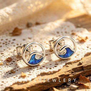 May include: A pair of silver-toned stud earrings featuring a circular frame with a stylized blue wave inlay. The wave design is filled with a vibrant, iridescent blue material that mimics the appearance of ocean water within the metal circle.