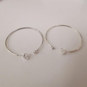 Puede incluir: Un par de pendientes de aro plateados. Cada pendiente tiene un diseño circular con una pequeña bola en un extremo y un poste con cierre de mariposa en el otro. Los pendientes se muestran sobre una superficie blanca.