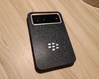 May include: A rectangular black smartphone with a textured finish and a prominent white BlackBerry logo in the center. The device features a horizontal camera module at the top with two lenses and a metallic copper-colored accent border.