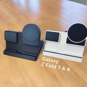May include: Two phone stands, one black and one white, designed for Galaxy Z Fold 7 & 8. Each stand features a circular charging pad and a rectangular pad with a carbon fiber texture. The stands are on a wooden surface.