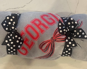 May include: Rolled gray blanket with the word "GEORGIA" in red block letters. The blanket is tied with two black bows with white polka dots and a red and black ribbon. The blanket is rolled up.