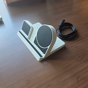 May include: A white multi-device charging station featuring a circular black magnetic charging pad and a rectangular black carbon fiber textured accent panel. The stand includes a black charging cable with a USB-C connector attached to the side.