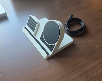 May include: A white multi-device charging station featuring a circular black magnetic charging pad and a rectangular black carbon fiber textured accent panel. The stand includes a black charging cable with a USB-C connector attached to the side.