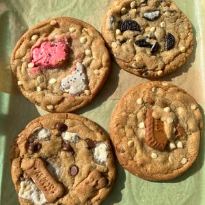 May include: Four gourmet cookies with various toppings are arranged on a green surface. One cookie has a pink frosted shape and white sprinkles. Another has Oreo cookie pieces. The third has marshmallows and chocolate chips. The last has a cookie with the word "Speculoos".