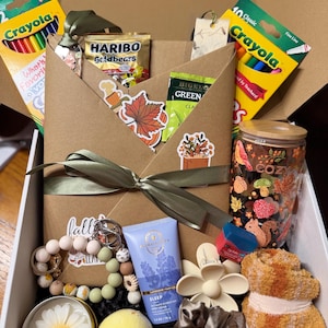 Puede incluir: Una caja de regalo que contiene un sobre marrón atado con una cinta verde, dos cajas de lápices de colores Crayola, una bolsa de Haribo Goldbears, una bolsita de té verde Bigelow, un vaso con motivos otoñales, un llavero de cuentas, crema corporal de lavanda, una pinza para el pelo en forma de flor, una bomba de baño amarilla, una vela de margarita, un coletero y calcetines suaves.