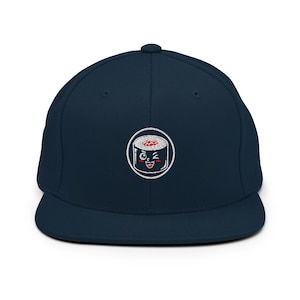 Peut inclure: Une casquette de baseball bleu marine avec un écusson circulaire brodé en blanc au centre. L'écusson représente un personnage de sushi en dessin animé avec un visage qui fait un clin d'œil, des joues rouges et un large sourire.