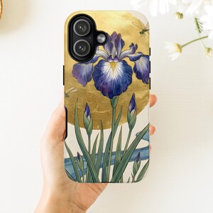 May include: A smartphone case featuring a large, detailed purple iris flower with green stems and buds set against a circular gold background. The design includes small dragonfly illustrations and a blue flowing line at the bottom, all on a white base.