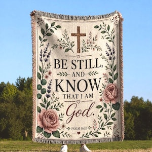 May include: A woven throw blanket featuring a cream background with a central brown cross and the text "BE STILL, AND KNOW THAT I AM GOD. PSALM 46:10". The design is framed by green foliage, lavender sprigs, and two large pink roses.