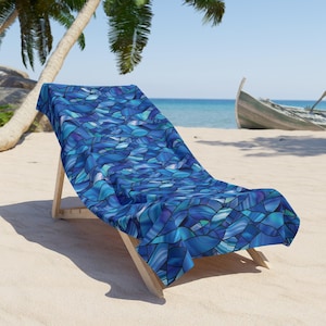 May include: A blue beach towel with a stained glass wave pattern is draped over a wooden beach chair. The towel's design features shades of blue, teal, and white. The scene is set on a sandy beach with a boat in the background.