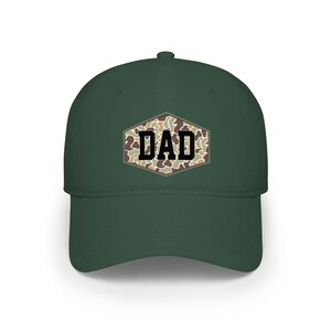May include: A dark green baseball cap with a curved peak. The cap displays the word "DAD" in black block letters on a camouflage patch. The camouflage patch has a brown, tan, and green pattern.