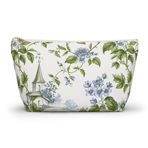 Op de afbeelding: Een witte make-up tas met een gouden rits, versierd met een bloemen- en kerkpatroon. Het patroon bevat groene bladeren, blauwe bloemen en een kleine kerk met een toren. De tas is rechthoekig en heeft een trapeziumvorm.