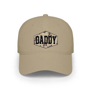 May include: A tan baseball cap with a curved brim. The front of the cap features the word "DADDY" in black block letters, set against a camouflage pattern within a hexagonal frame. The cap is a classic six-panel design.
