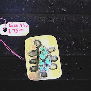 May include: A rectangular, gold-toned pendant with a turquoise stone center. The pendant has a unique, openwork design with dark gray accents. A tag with the text "Size 4 1/2" is attached with pink string.
