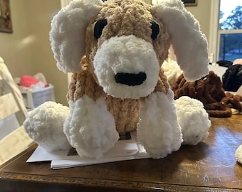 Puede incluir: Un juguete de perro de peluche hecho a mano, con una combinación de colores marrón y blanco. El perro tiene orejas blancas grandes y caídas, un hocico blanco y ojos negros. El juguete está sobre una mesa de madera.