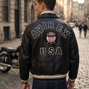 May include: Black leather jacket with white lettering and patches. The back of the jacket features the word "AVIREX" above a patch with the words "All Americans" and "USA". The jacket has a cream-colored collar and cuffs.