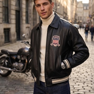 May include: A black leather bomber jacket with a cream-colored turtleneck and dark blue jeans. The jacket features a patch with red, white, and blue colors and the text "AVIREX ESTABLISHED 1975". A motorcycle is visible in the background.