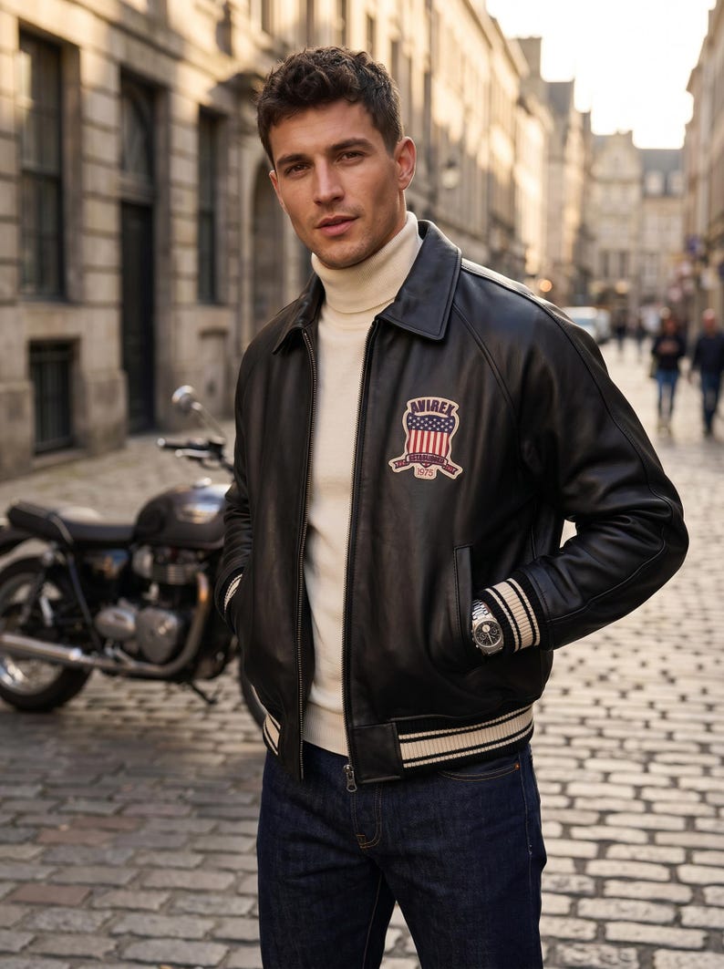 May include: A black leather bomber jacket with a cream-colored turtleneck and dark blue jeans. The jacket features a patch with red, white, and blue colors and the text "AVIREX ESTABLISHED 1975". A motorcycle is visible in the background.