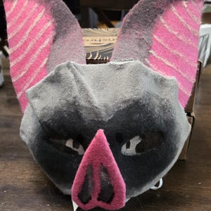 May include: A handmade bat mask with large pink and black ears, a gray face, and a pink nose. The mask has mesh eye coverings and is likely for costume or cosplay purposes.