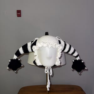 May include: A whimsical, handmade crochet hat in black and white stripes, featuring a white ruffled brim and black claw-like embellishments. The hat is displayed on a mannequin head, showcasing its unique design.