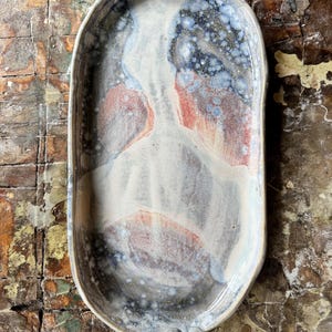 Puede incluir: Bandeja de cerámica hecha a mano con una forma orgánica. La bandeja presenta una mezcla de colores, blanco, azul y rojo, con una superficie moteada y texturizada. Tiene tres pequeños pies redondeados en cada extremo. Una pieza decorativa.