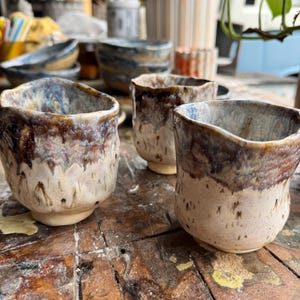 Puede incluir: Tres tazas de cerámica hechas a mano con un esmalte texturizado único. Las tazas presentan una base beige claro con un borde marrón oscuro y azul. Las tazas están sobre una superficie de madera rústica.