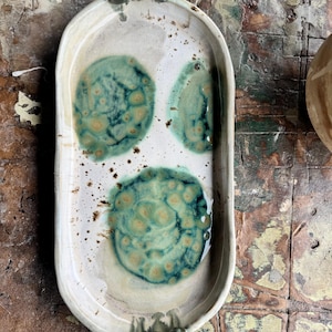 Puede incluir: Una bandeja de cerámica hecha a mano con una base de color crema y tres diseños circulares en verde azulado y amarillo. La bandeja rectangular tiene bordes redondeados y pequeños detalles decorativos de color gris oscuro en cada extremo.