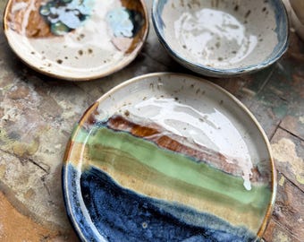 May include: A collection of handmade ceramic tableware. The set includes two small plates and a bowl. The plates feature unique designs with blue, green, brown, and white glaze. The bowl has a blue rim and a speckled interior. The pieces are on a wooden surface.