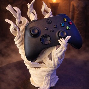 May include: A black Xbox controller with colorful buttons is held by a white, wrapped hand sculpture. The background features an ancient Egyptian theme with statues and a torch.