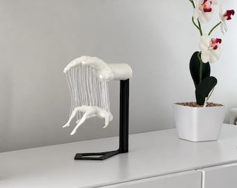May include: A white sculpture of a hand suspended above a small white human figure, connected by thin white strings. The sculpture is mounted on a black L-shaped stand. The hand is positioned horizontally, with the figure hanging below it.