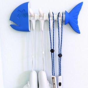 May include: A blue and white fish-shaped wall organizer with a cross design on the head and tail fin. It holds white game controllers and blue and white lanyards with black accents. A fun and functional storage solution.