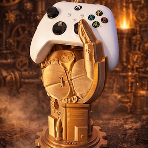 May include: A white Xbox controller sits atop a gold-coloured, steampunk-style hand-shaped stand. The stand has intricate mechanical details and a gear-shaped base, against a blurred background of gears and machinery.