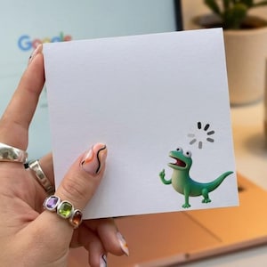 May include: A square white note card featuring a small green lizard illustration in the bottom right corner, positioned below a circular loading icon. The card is held against a blurred background showing a laptop, a potted plant, and a coffee mug.