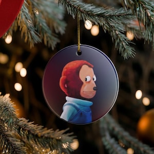 May include: A round Christmas ornament featuring a cartoon monkey with red fur and a blue outfit. The ornament hangs from a gold string against a backdrop of a decorated Christmas tree with red ribbon and other ornaments.