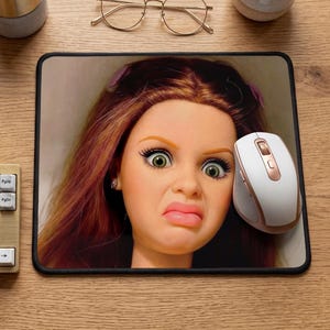 May include: A rectangular mousepad featuring a printed graphic of a doll face with long brown hair, wide green eyes, and a frowning expression. The mousepad is positioned on a wooden desk next to a white and rose gold computer mouse, a keyboard, a notebook, a pen holder, and glasses.