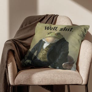 May include: A decorative throw pillow featuring a frog dressed in formal attire. The frog is wearing a black coat and vest, with the text "Well, shit." printed above. The pillow is placed on a beige chair with a brown blanket draped over it.