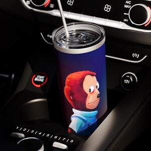 May include: A stainless steel tumbler with a blue background and a cartoon monkey illustration. The tumbler has a silver lid and a silver straw. The tumbler is in a car's cup holder.